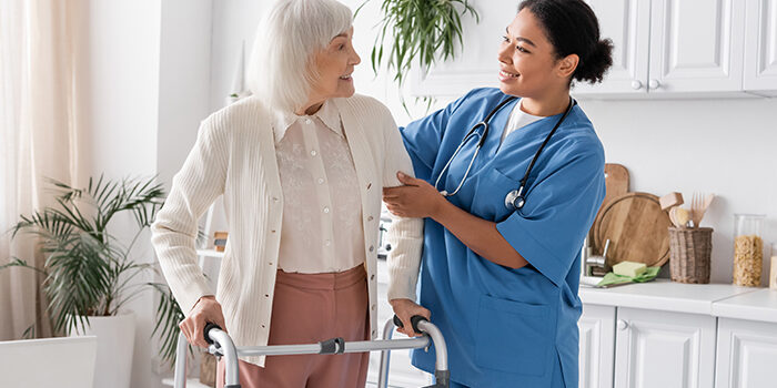 happy multiracial nurse in blue uniform supporting senior woman with grey hair walking with help of walker,stock image